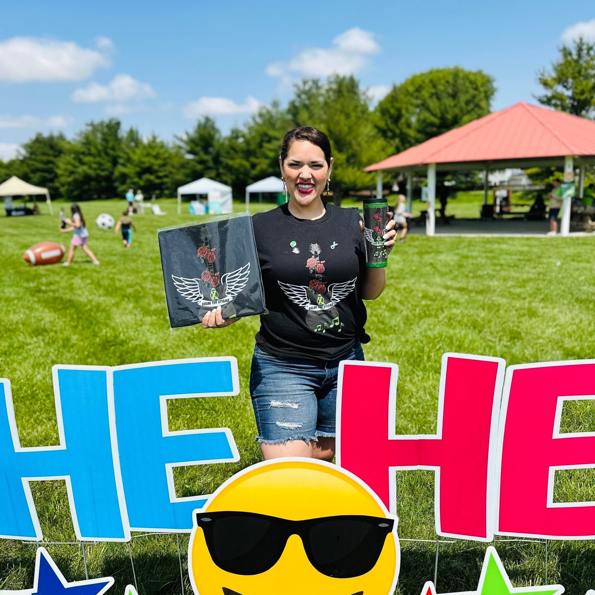 Miriam at an outdoor community event, holding a Break the Stigma shirt and tumbler